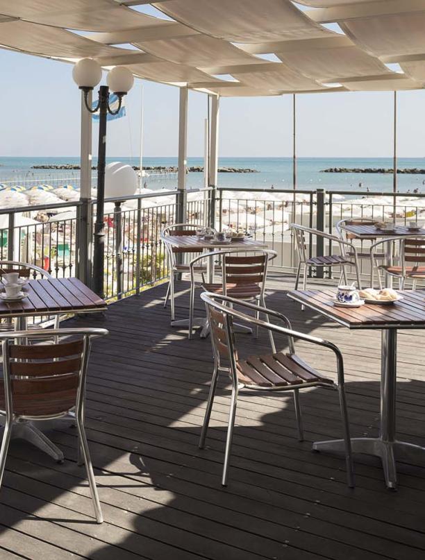 Terrazza con vista mare, tavoli e sedie, spiaggia e ombrelloni.